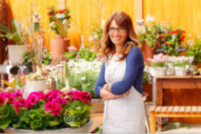 Consultanta fonduri UE pentru femei - finantare europeana. O doamna proprietara de florarie sta zambind in fata magazinului, iar in background se vad flori multicolore.